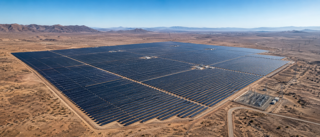 Texte alternatif : Vue aérienne panoramique d'une centrale solaire photovoltaïque de 100 MW installée dans une vaste plaine désertique au Maroc, avec un poste de raccordement électrique au premier plan et des montagnes en arrière-plan.