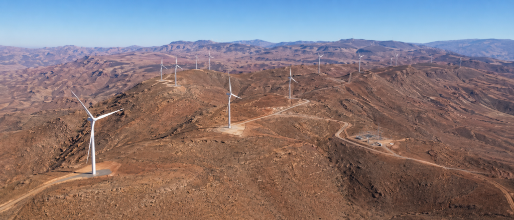 Vue aérienne d'un parc éolien de 100 MW installé sur les crêtes d'une montagne aride dans le sud du Maroc, montrant l'alignement des turbines le long des pistes d'accès techniques sous un ciel dégagé.